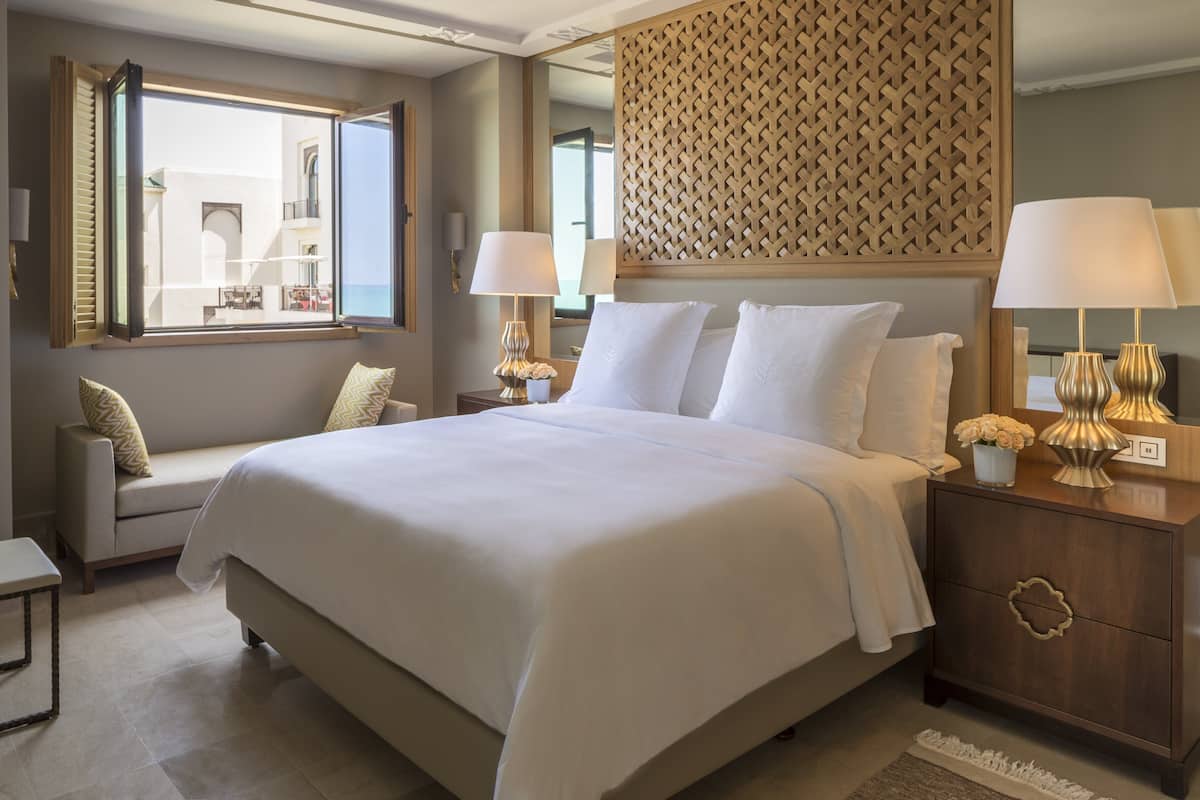 Quarto de hotel com uma cama grande, área de estar com um banco, mesa de cabeceira com luminárias e vista pela janela.