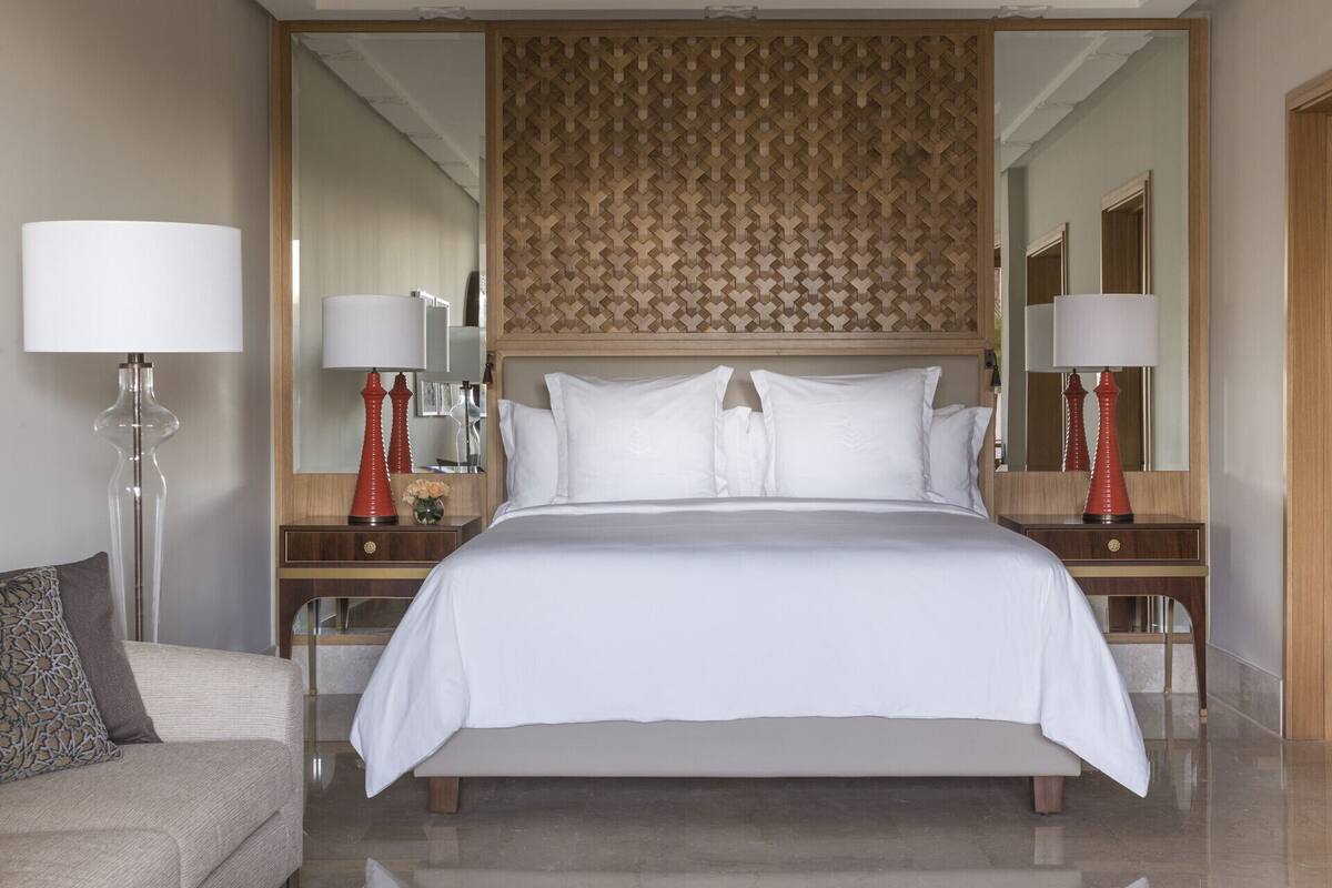 Quarto de hotel com uma cama grande, duas mesas de cabeceira com luminárias, um espelho e um painel de cabeceira estampado.