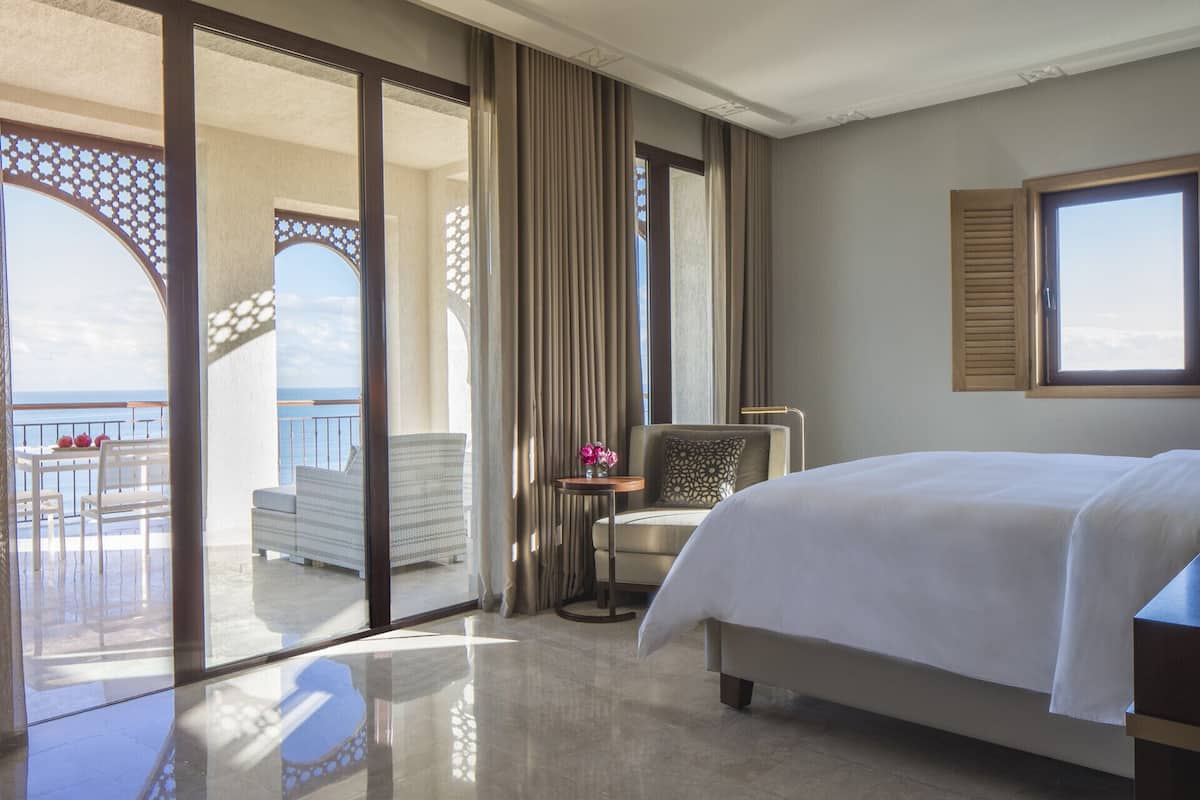 Quarto de hotel com cama, uma cadeira, uma mesinha com flores, vista para o mar e varanda com mobiliário para área externa.