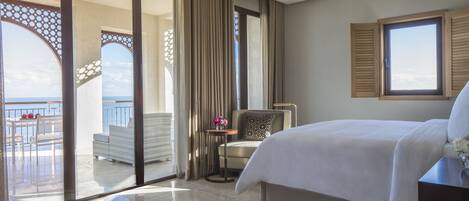 A hotel room with a bed, a chair, a small table with flowers, a view of the sea, and a balcony with outdoor seating.