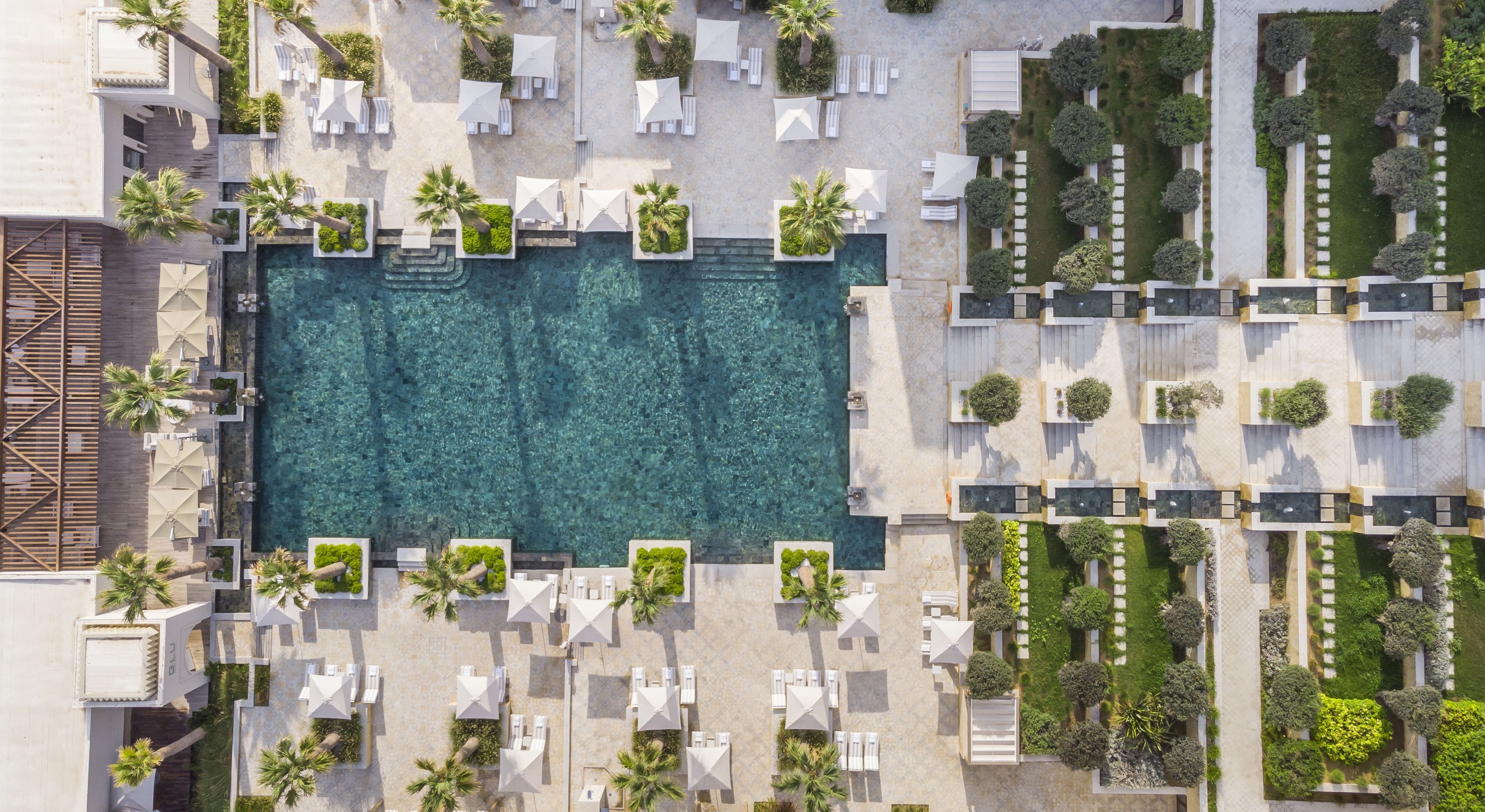 An aerial view of a resort with a central swimming pool, surrounded by lounge areas with white umbrellas and chairs, and a variety of landscaped greenery.