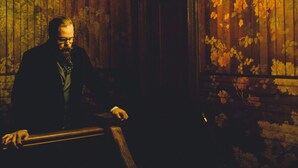 Un hombre con traje oscuro sentado junto a una barandilla de madera, con un fondo de papel tapiz floral y una araña colgante.