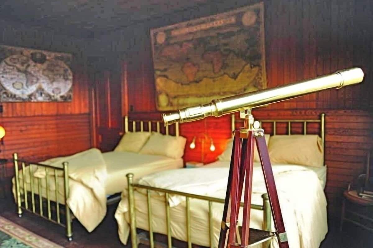 Una habitación con un telescopio sobre un trípode, dos camas, un mapa en la pared y una lámpara.