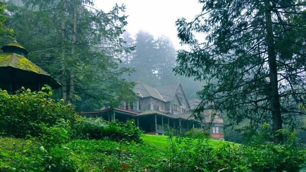 Una casa grande de dos pisos con un techo empinado, rodeada de un bosque denso.