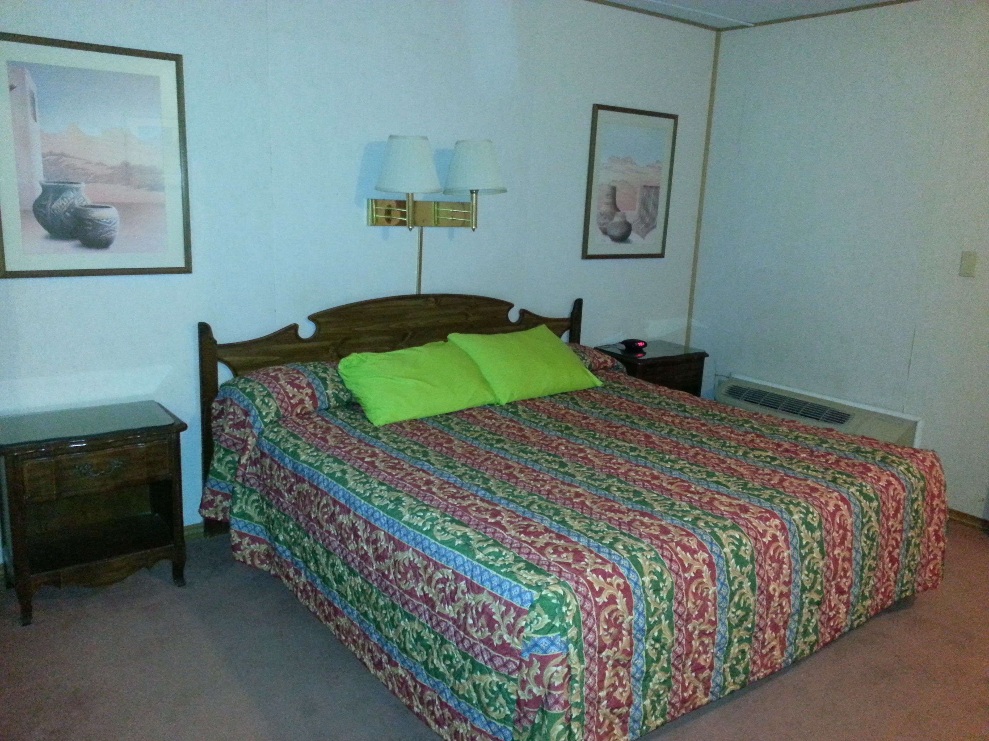 Un dormitorio con una cama de madera, almohadas verdes, una colcha estampada, una mesita de noche, una lámpara y dos cuadros en la pared.
