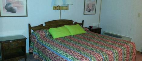 Un dormitorio con una cama de madera, almohadas verdes, una colcha estampada, una mesita de noche, una lámpara y dos cuadros en la pared.