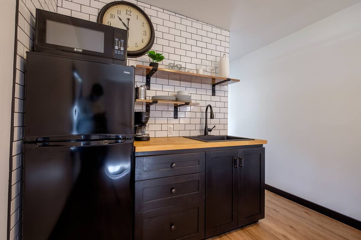 A compact kitchen with a black refrigerator, a microwave, a clock, and a wooden countertop.