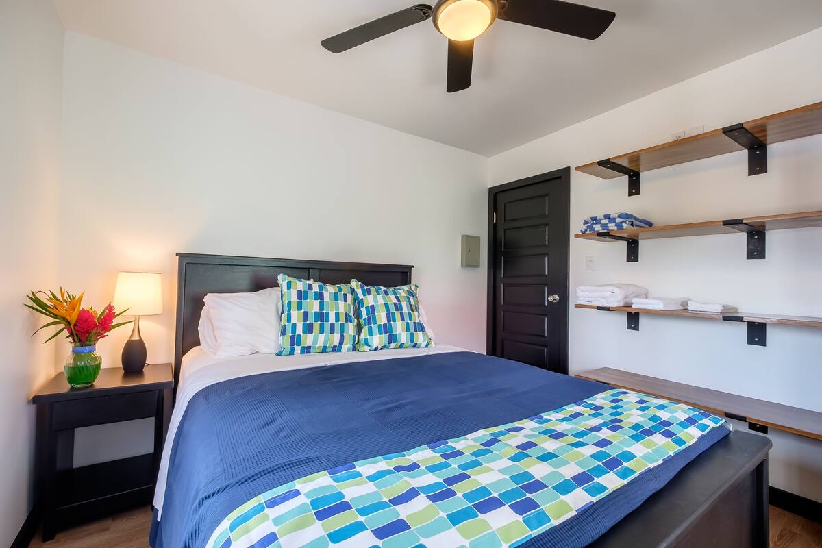 A neatly made bed with a blue and white checkered bedspread, a dark headboard, and two pillows with a checkered pattern. There is a nightstand with a lamp and a vase of flowers to the left. A ceiling fan is visible above the bed.
