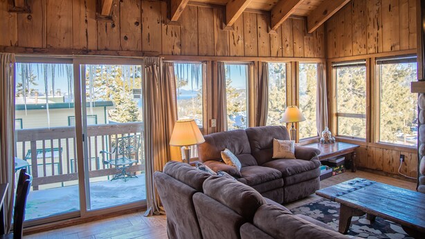 A cozy wooden interior with a sectional sofa, a coffee table, and a lamp. Large windows offer a view of the outdoors. - Lake Views! Perfect Mountain Summer Get-a-way No traffic, 8,000 ft. elevation (Stateline)