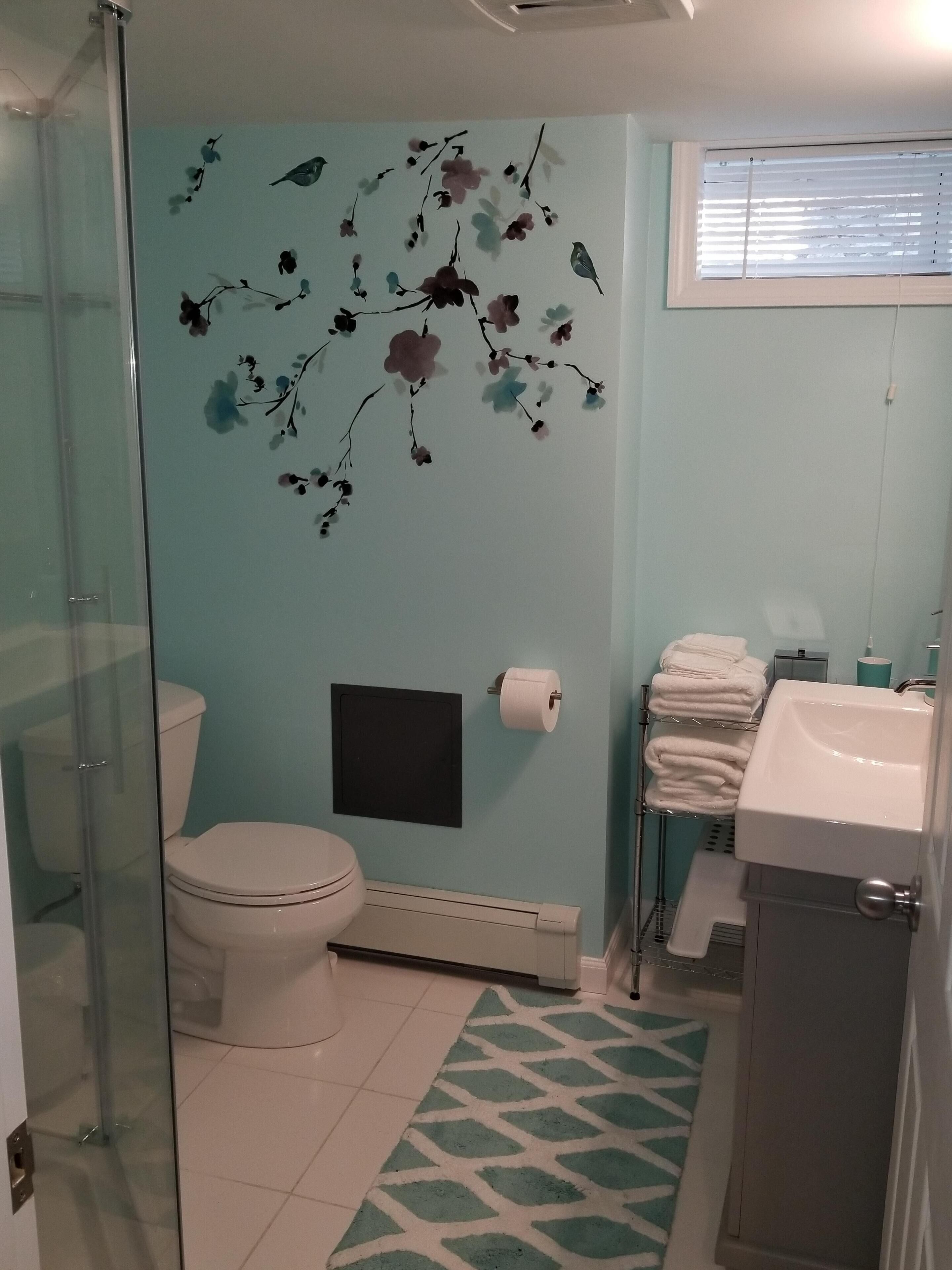 Een badkamer met toilet, wastafel en douche. Er is een handdoekrek met handdoeken, een toiletpapierhouder en een decoratieve muur met bloemmotieven.