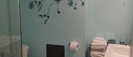 Een badkamer met toilet, wastafel en douche. Er is een handdoekrek met handdoeken, een toiletpapierhouder en een decoratieve muur met bloemmotieven.