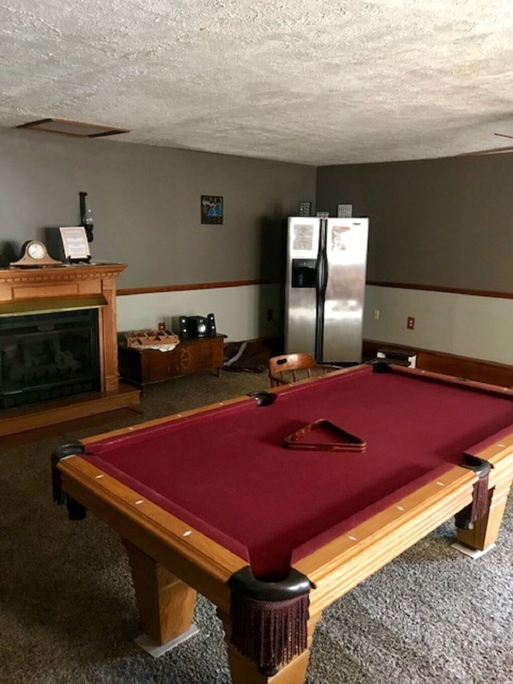 Une table de billard recouverte d’un feutre rouge, une cheminée avec une manteau de cheminée, et un réfrigérateur dans une pièce dont le sol est recouvert de moquette et dont les boiseries sont en bois.