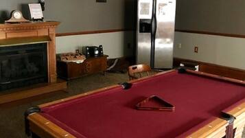 Une table de billard recouverte d’un feutre rouge, une cheminée avec une manteau de cheminée, et un réfrigérateur dans une pièce dont le sol est recouvert de moquette et dont les boiseries sont en bois.