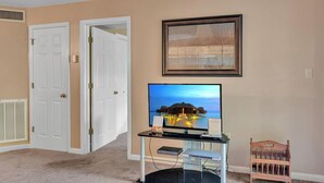 Ein Zimmer mit beigen Wänden, einem Fernseher auf einem Glastisch, einem gerahmten Bild über dem Fernseher und einem hölzernen Babybett auf der rechten Seite.