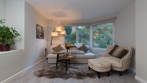 Ein gemütliches Zimmer mit Sofa, Sessel, Lampe, Pflanze und einem Fenster mit Blick auf Grünflächen.