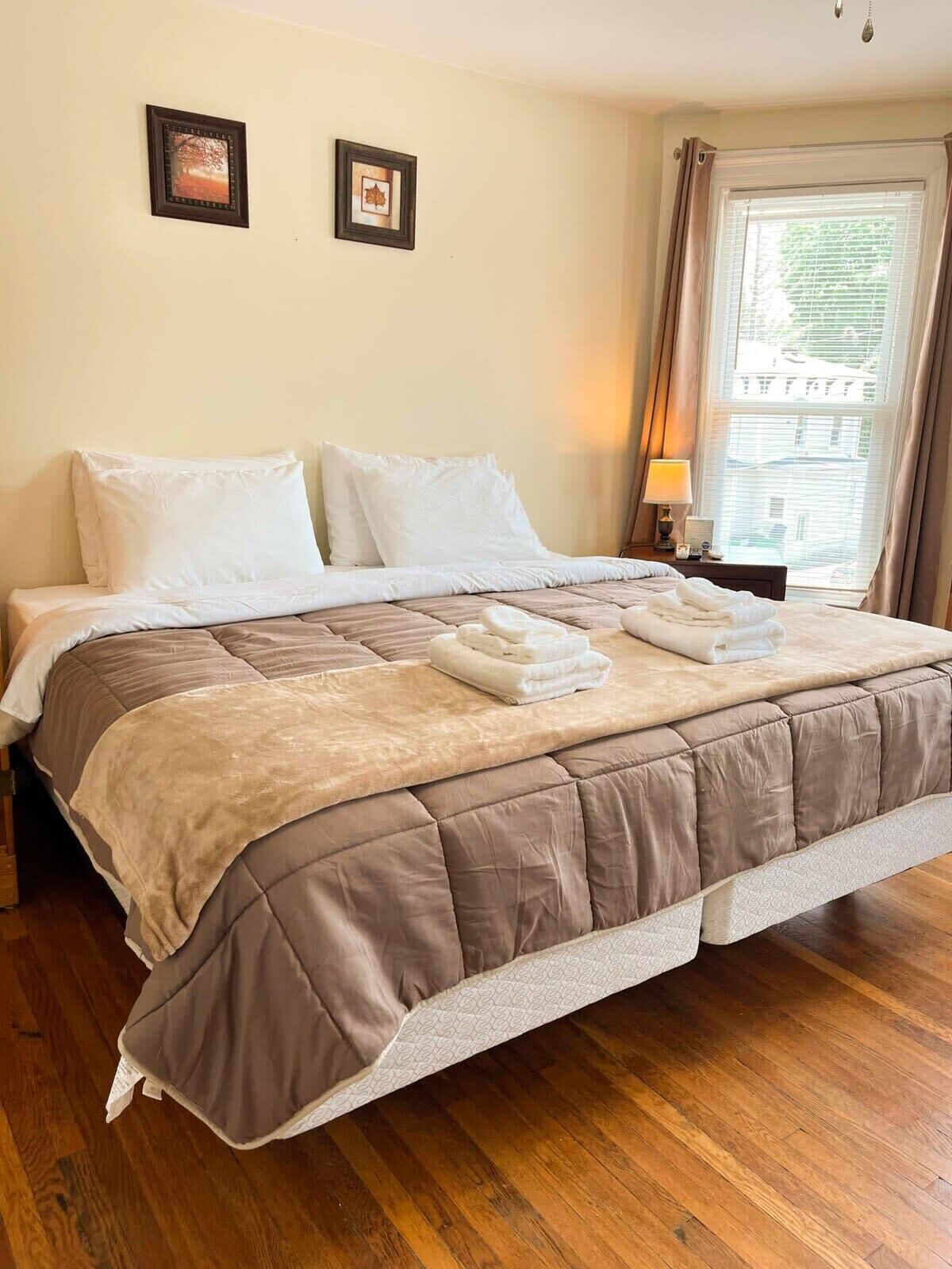 A neatly made bed with a brown quilt, white pillows, and folded towels on a wooden floor.