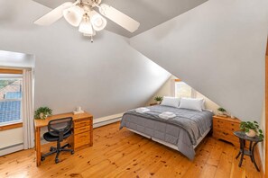 A bedroom with a sloped ceiling, a bed with a grey duvet, a wooden desk with a chair, and a small table with a plant. - After the Game Apt 1 - All Star Baseball Rentals (Oneonta)