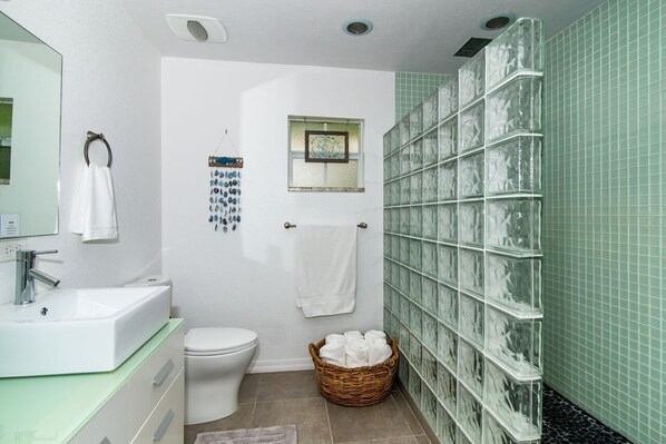 Un baño con lavamanos blanco, inodoro, espejo, toallero, cesta de toallas y cabina de ducha de vidrio.