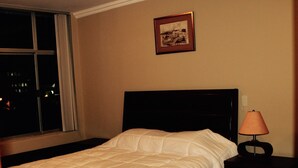 A neatly made bed with a white comforter in a room with a window, a framed picture on the wall, and a bedside lamp.