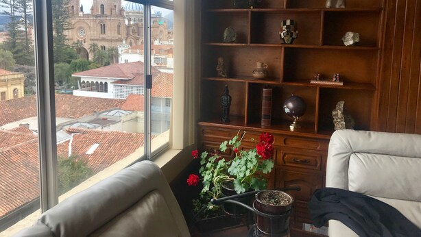 A room with a large window overlooking a historic building with a dome and arches. Inside, there is a wooden bookshelf with various decorative items and a potted plant with red flowers.