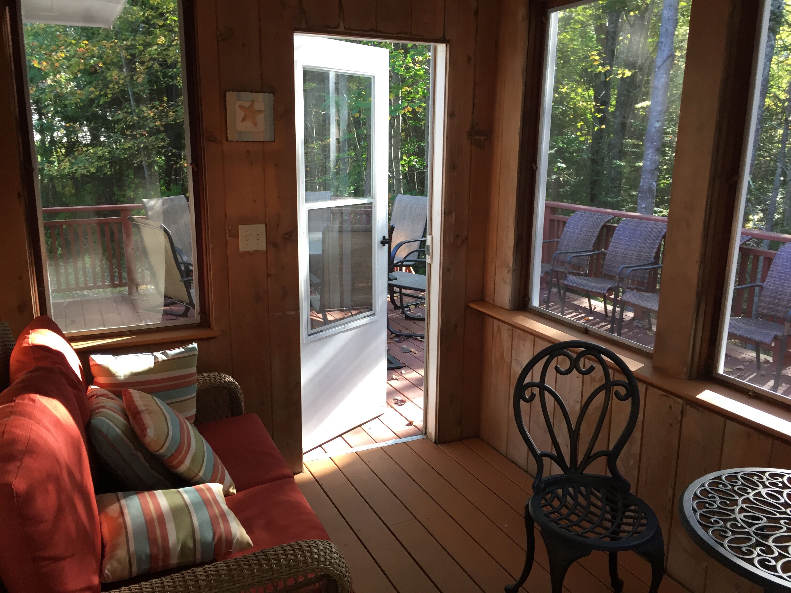 A wooden deck with a red sofa and a wicker chair, a small round table, and a view of a deck chair area through a glass door.