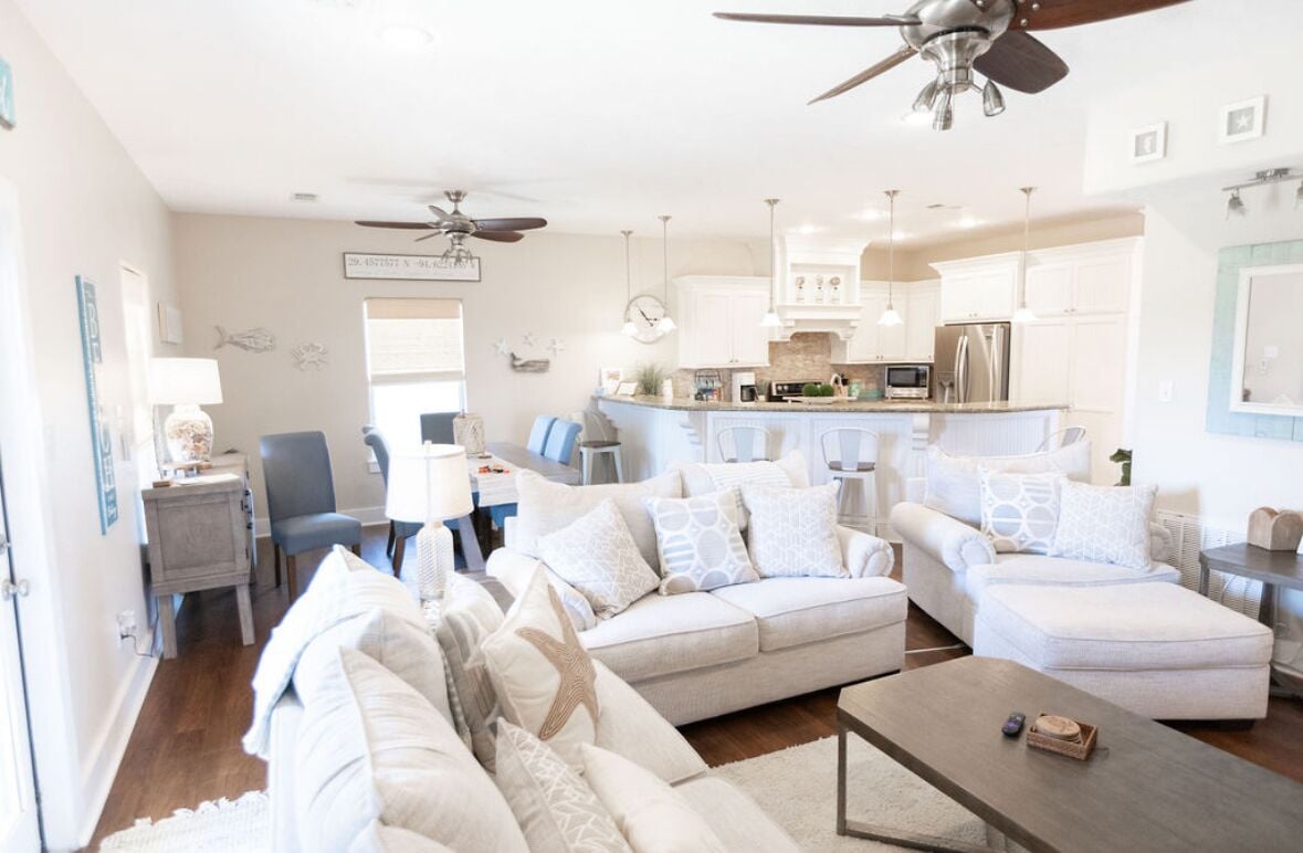 A spacious living room with a large sofa, a coffee table, and a ceiling fan. There is a kitchen area with white cabinets and a dining table.