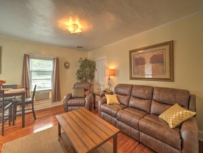 A living room with a brown leather sofa, a wooden coffee table, a dining table with chairs, and a window with curtains.