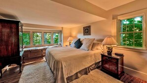 Quarto com uma cama grande, uma mesa de cabeceira de madeira, um banquinho e uma janela com vista para o verde.