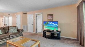 Ein Wohnzimmer mit braunem Ledersofa, einem Glastisch, einem Flachbildfernseher auf einem schwarzen Standfuß, beige gefärbten Wänden und einer Tür im Hintergrund.