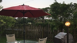 En uteplats med ett glasbord, två stolar, ett rött parasoll och en grillplats under tak.