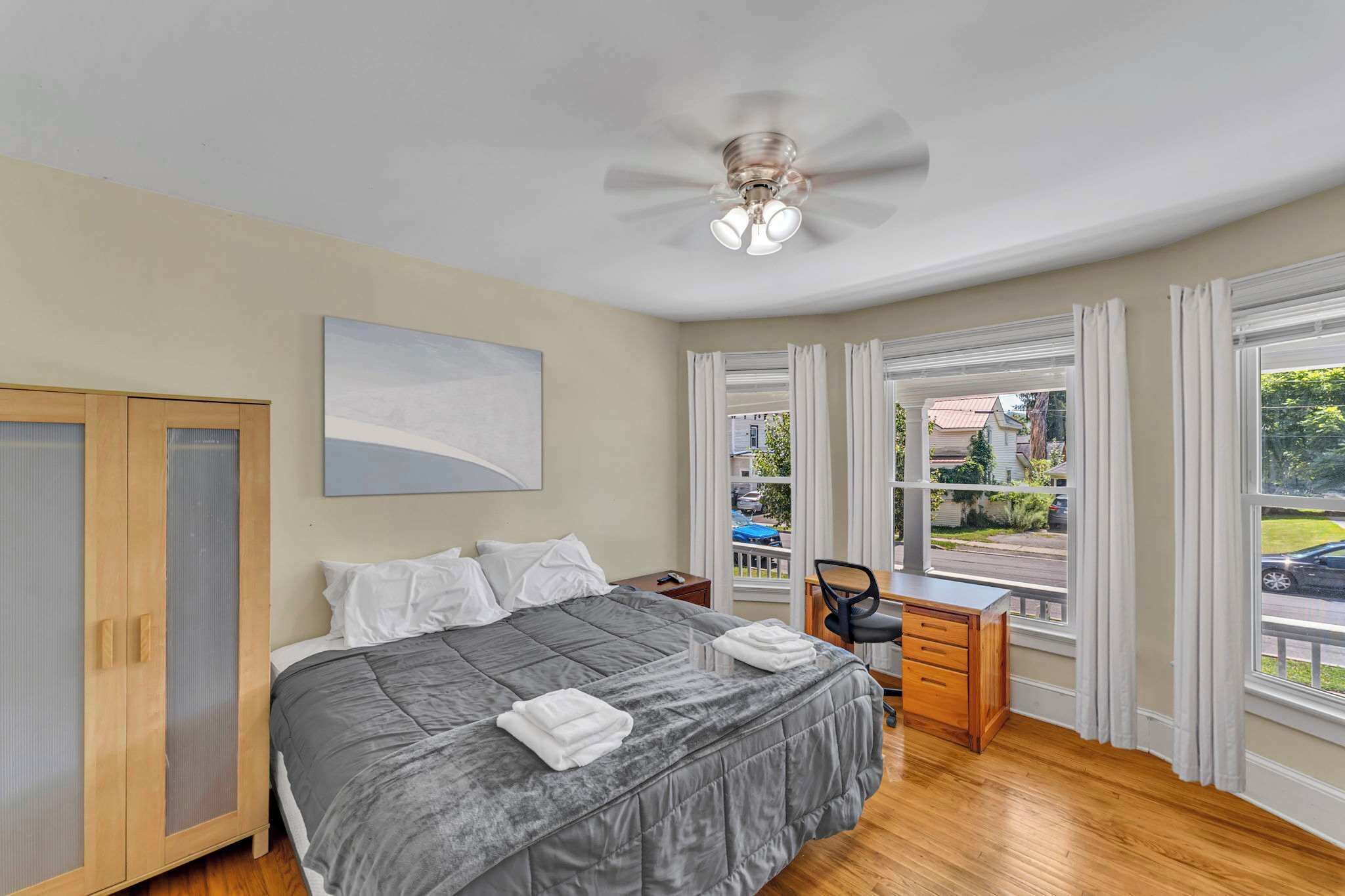 A bedroom with a bed, a desk, and a view of the street through large windows.
