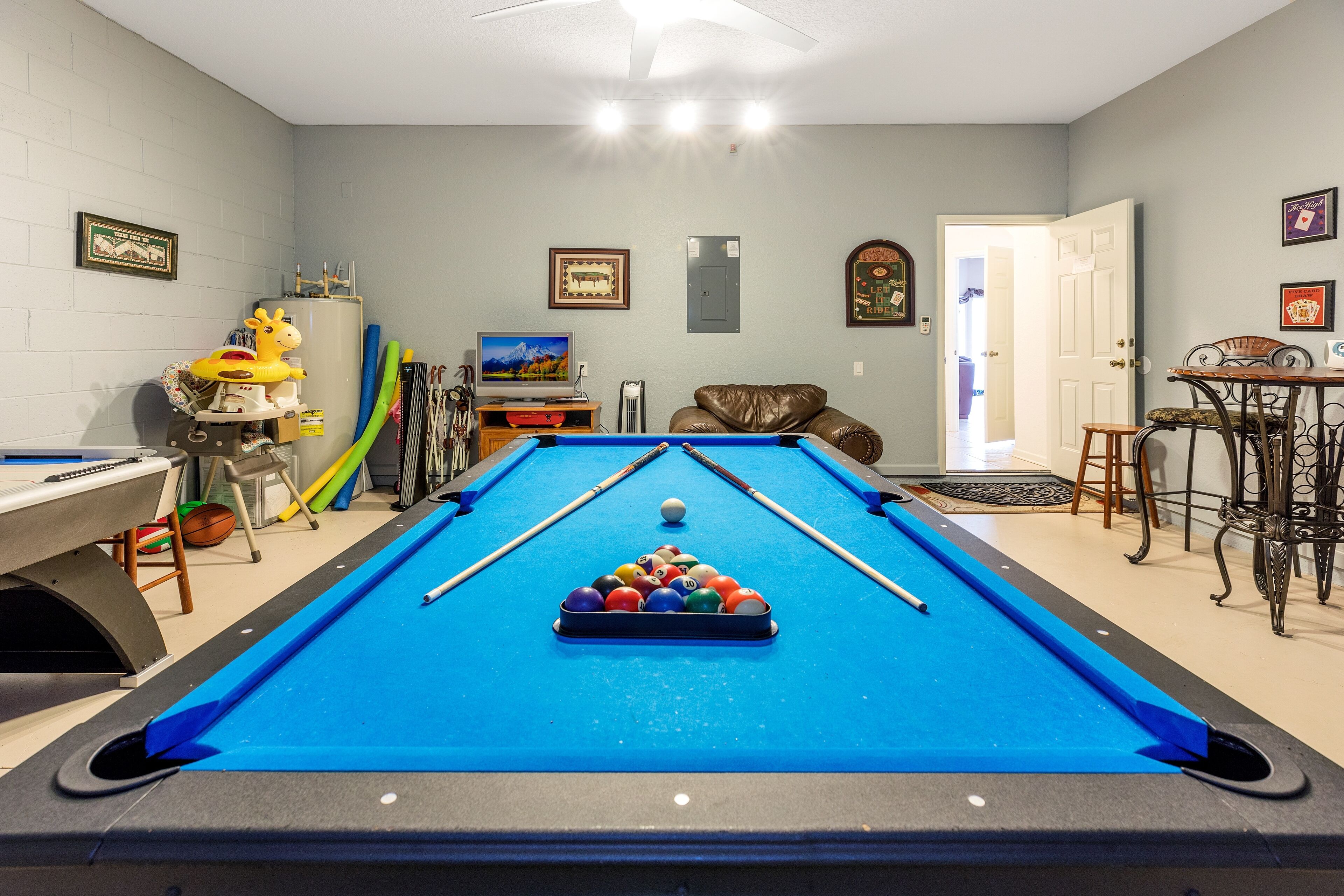 A pool table with a blue felt surface, surrounded by a brown leather couch, a television, and various pool cues.