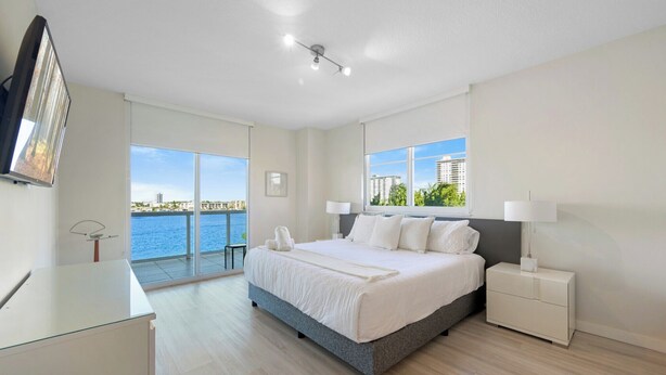 A modern bedroom with a large bed, a nightstand, a desk, and a view of the water and buildings. - Luminous corner condo overlooking the bay | WIFI + Netflix + Parking (Sunny Isles Beach)