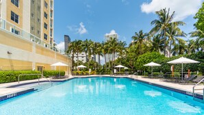 A swimming pool with lounge chairs and umbrellas, surrounded by palm trees and a building with balconies. - Luminous corner condo overlooking the bay | WIFI + Netflix + Parking (Sunny Isles Beach)