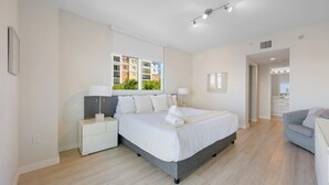 A modern hotel room with a large bed, bedside tables, a window with a view of buildings, and a separate seating area with a chair. - Luminous corner condo overlooking the bay | WIFI + Netflix + Parking (Sunny Isles Beach)
