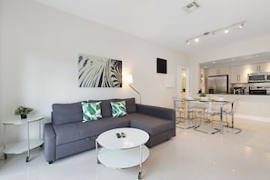 Uma sala de estar moderna com um sofá cinza, uma mesa de centro redonda e uma área de jantar com mesa e cadeiras de vidro.