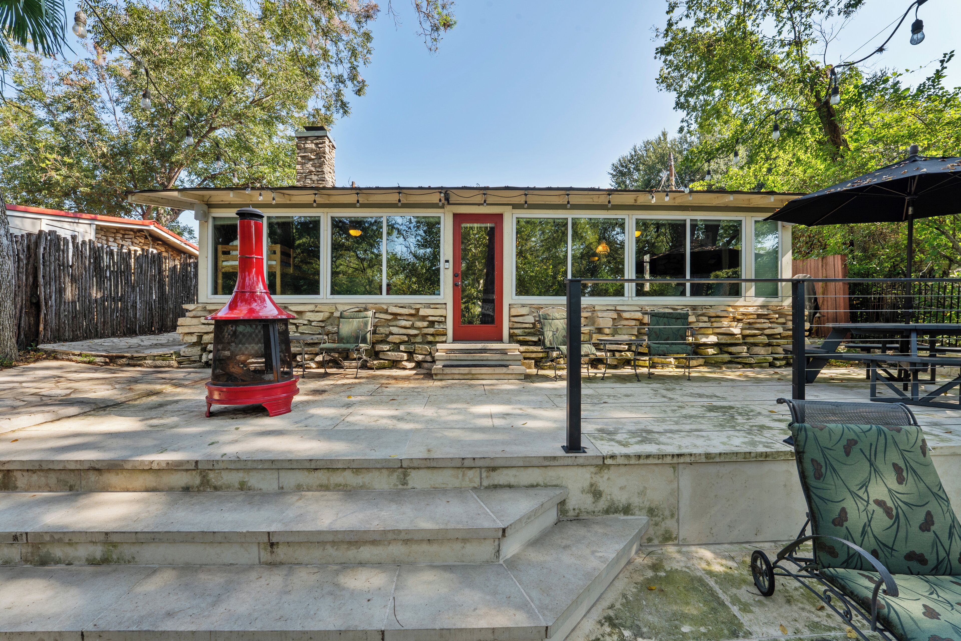 Une maison en pierre avec une porte rouge, une cheminée et une terrasse aménagée avec du mobilier d’extérieur.