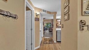 A hallway with a wooden floor, a door on the left, and a kitchen area with a countertop and cabinets visible in the background.