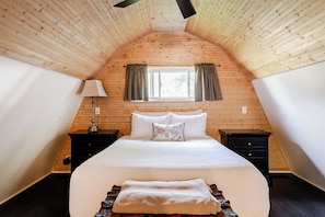 A bedroom with a sloped ceiling, a bed with white bedding, a nightstand, a lamp, and a window with curtains.