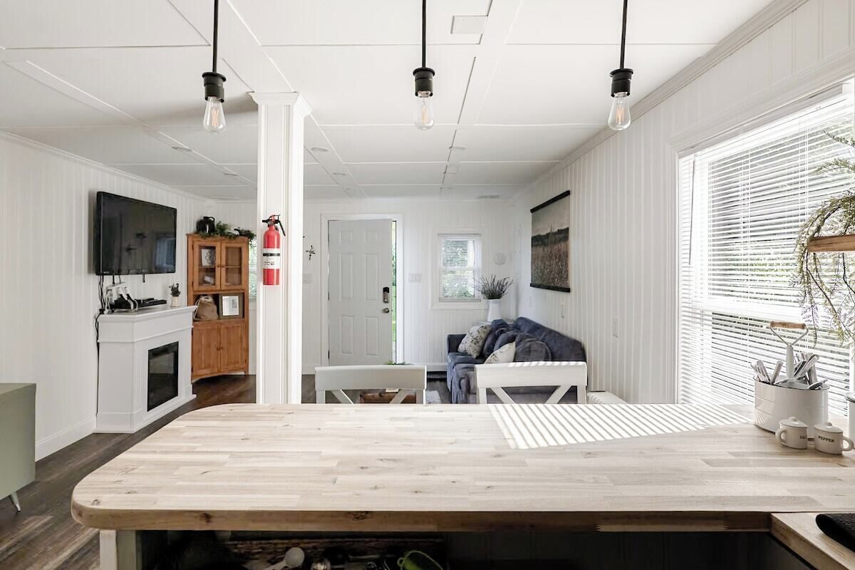 Une cuisine moderne avec un îlot central en bois, des meubles blancs et un four encastré. On y trouve un coin repas avec une table et des chaises blanches, ainsi qu’une grande fenêtre équipée de stores.