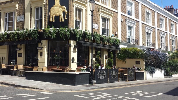 A corner building with a sign reading "CASTLE" and "ELEPHANT & CASTLE" on the ground floor. There are outdoor seating areas and a street sign indicating "GORDON PLACE INN."