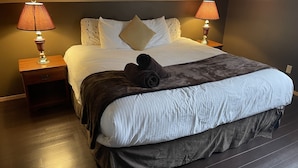 A neatly made bed with a white duvet, a dark throw blanket, and a rolled towel. There are two bedside lamps on wooden nightstands. - VACATION RENTAL- SLEEPS UP TO 10 -GOLDEN BC (Blaeberry)