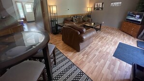 A living room with a brown leather sofa, a glass coffee table, and a dining table with chairs. There is a TV on a wooden cabinet and a floor lamp. - VACATION RENTAL- SLEEPS UP TO 10 -GOLDEN BC (Blaeberry)