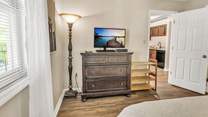 Une chambre à coucher avec un lit, une commode, une lampe, un téléviseur et une vue sur la cuisine par une porte ouverte.