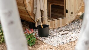 Eine Holzsauna mit geöffneter Tür, ein Handtuch am Türhänger, ein Eimer und ein Paar Hausschuhe auf dem Boden.
