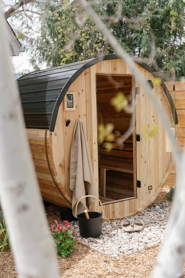 Eine Holzsauna mit geöffneter Tür, ein Handtuch am Türhänger, ein Eimer und ein Paar Hausschuhe auf dem Boden.