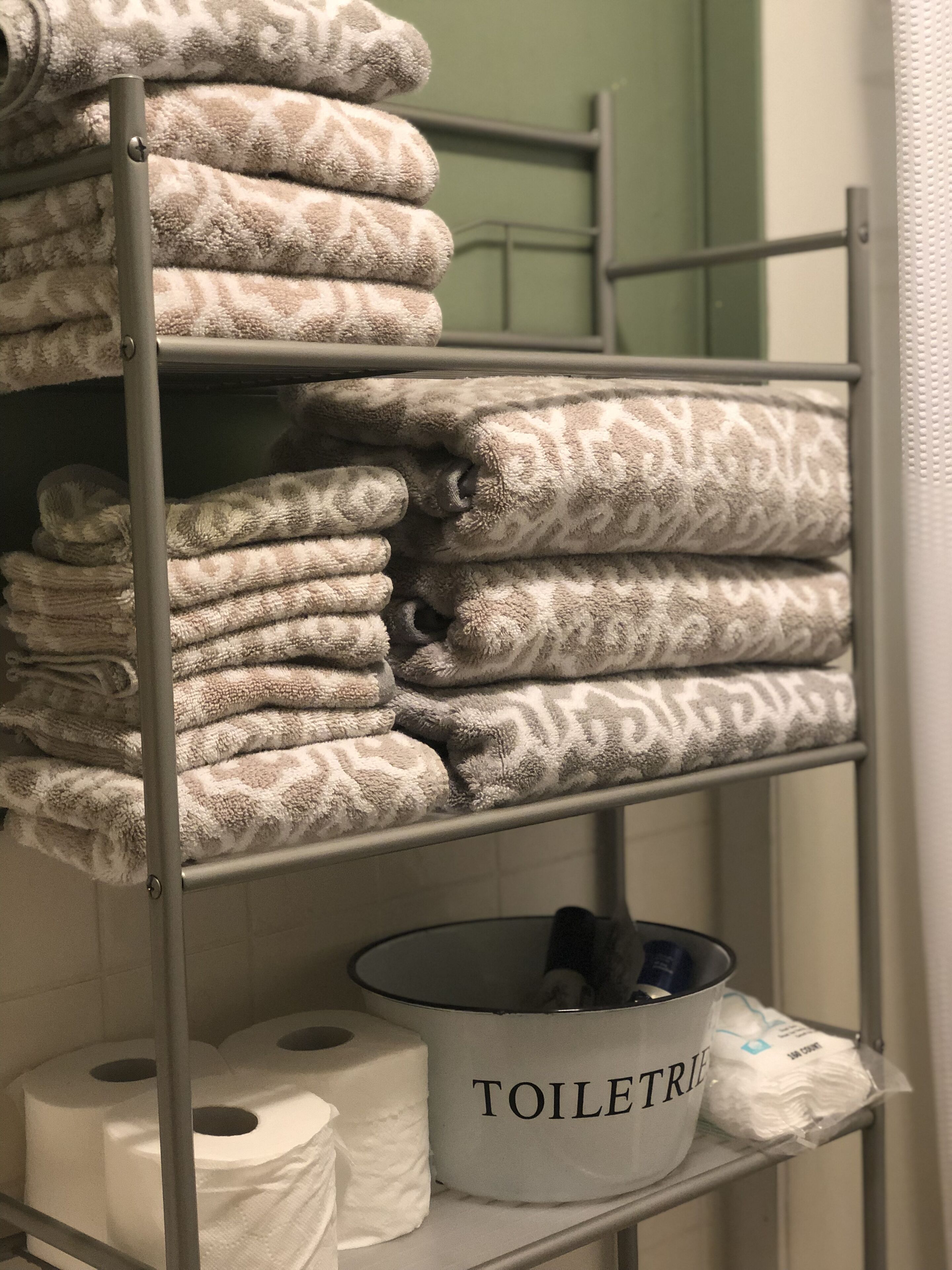 A bathroom with a shelf holding towels and a container labeled "TOILET" with toilet paper and other toiletries.