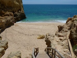 Una playa con una escalera de madera que baja hacia la orilla, rodeada de acantilados rocosos.