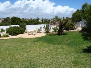 Un patio trasero con césped, una mesa y sillas blancas, rodeado de plantas variadas y un muro blanco.