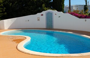 Una piscina de borde curvo, con un solárium de baldosas y una pared blanca con una puerta decorativa.